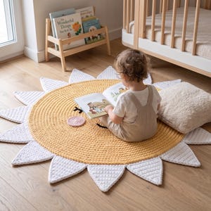 Puede incluir: Alfombra de ganchillo amarilla y blanca con forma de sol. La alfombra tiene un centro amarillo con una cara sonriente y rayos triangulares blancos. Un ni&ntilde;o est&aacute; sentado en la alfombra, leyendo un libro. Una peque&ntilde;a posavasos rosa est&aacute; en la alfombra.