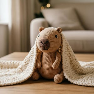 Pu&ograve; includere: Un orsetto all'uncinetto fatto a mano, nei toni del marrone, coperto da una coperta strutturata. L'orso ha un corpo rotondo, piccole orecchie e un muso dettagliato. La coperta &egrave; color crema. L'orsetto &egrave; seduto su una superficie di legno.