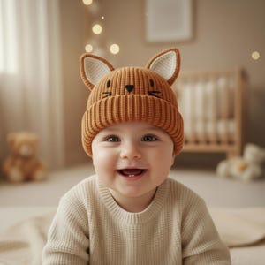 Pode incluir: Um beb&ecirc; sorrindo usando um gorro de malha marrom com orelhas de raposa e um desenho de rosto preto. O beb&ecirc; tamb&eacute;m est&aacute; usando um su&eacute;ter creme. O fundo inclui um ber&ccedil;o e um ursinho de pel&uacute;cia.