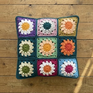 May include: A square crochet pillow with a dark teal border and nine colorful flower squares. Each square features a different color flower, including white, yellow, orange, and pink, on a variety of colored backgrounds. The pillow is placed on a wooden surface.