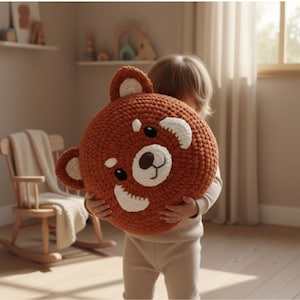 Pode incluir: Uma almofada grande e redonda de crochê em forma de panda vermelho. A almofada é principalmente laranja com detalhes brancos ao redor dos olhos e orelhas. A almofada de panda vermelho está sendo segurada por uma pessoa.
