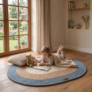 May include: A round braided rug with blue, tan, and cream concentric circles. A child is lying on the rug, reading a book, with a teddy bear and a woven pillow nearby. The rug is on a wooden floor.