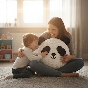 May include: A large, round, white and black panda bear pillow. The pillow has black ears, eyes, and a nose, with a smiling face. The pillow is being held by a person and a child.