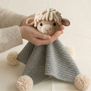 May include: A handmade crochet lamb security blanket. The lamb's head is beige with a textured, cream-colored mane and small, rounded ears. The blanket is gray with four cream-colored pom-poms at the corners. The lamb is held in two hands.