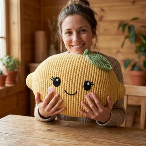 May include: A crocheted lemon-shaped pillow in yellow with a green leaf. The pillow has black eyes, eyelashes, and a smiling mouth. Pink blush is on the cheeks. The pillow is held by a person.