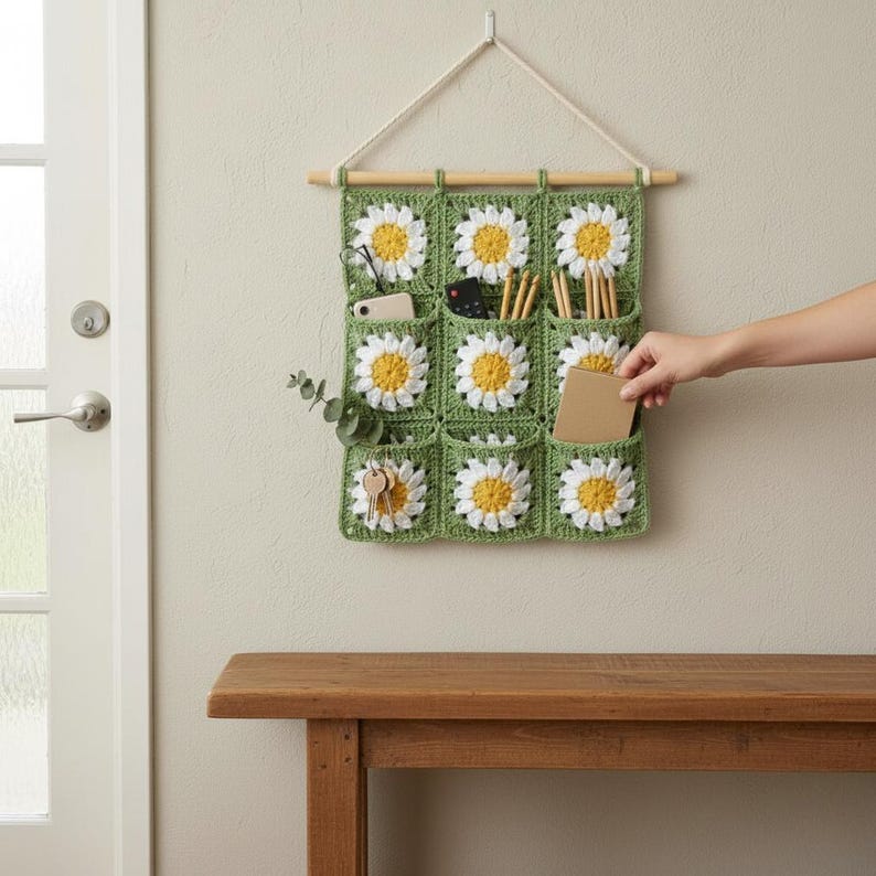 May include: A handmade, green crochet wall organizer with nine pockets, each adorned with a daisy motif. The organizer is suspended from a wooden dowel and a white cord. The pockets hold items like keys, pencils, and a small notebook.
