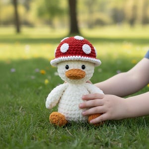 May include: A handmade crochet duck wearing a red mushroom hat with white spots. The duck has an orange beak and feet, and is sitting on green grass. The duck is a whimsical, soft toy.