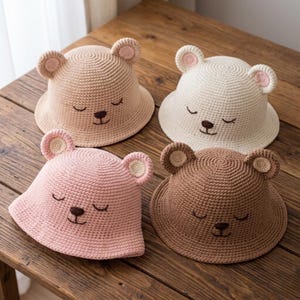 May include: Four crochet bear hats in beige, white, pink, and brown. Each hat features bear ears and a stitched face with closed eyes and a small nose. The hats are displayed on a wooden surface.