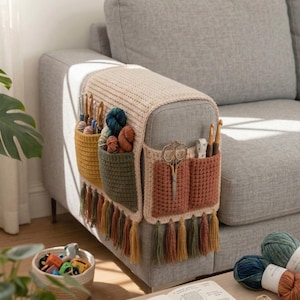 K&ouml;nnte beinhalten: Ein beigefarbener, senfgelber und rostfarbener H&auml;kel-Sofa-Arm-Organizer mit mehreren Taschen und bunten Quasten. Der Organizer ist mit H&auml;kelnadeln, Garn, Scheren und einem Ma&szlig;band gef&uuml;llt. Der Organizer ist am Arm eines grauen Sofas befestigt.