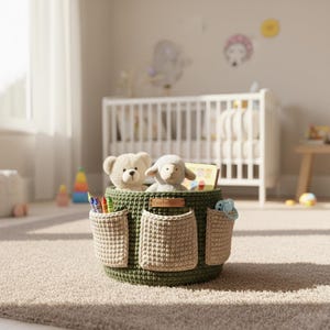May include: A green crochet storage basket with three beige pockets, filled with toys and crayons. The basket contains a teddy bear, a lamb, and a book. The basket is sitting on a light brown carpet.