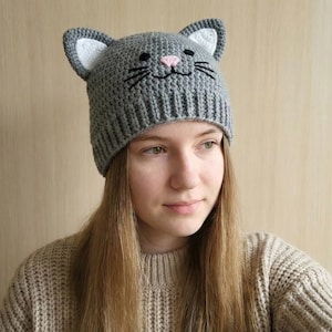 Könnte beinhalten: Graue gehäkelte Katzenmütze mit weißen Innenohren, einer rosa Nase und schwarzen gestickten Schnurrhaaren. Die Mütze ist im Beanie-Stil und wird von einer Person mit langen blonden Haaren getragen. Ein lustiges und skurriles Accessoire.