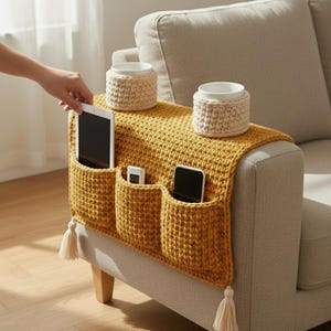 May include: Mustard yellow crochet sofa armrest organiser with three pockets holding a tablet, a remote, and a smartphone. Two beige crochet cup holders sit on top. The organiser has cream-coloured tassels on the ends.