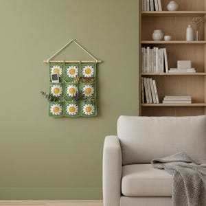 May include: A green crochet wall organizer with nine pockets, each featuring a daisy design with yellow centers and white petals. The organizer hangs from a wooden dowel and holds a phone, glasses, and small plants. The background is a light green wall.