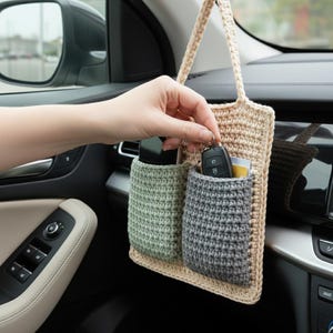May include: A beige crochet car organizer with two pockets in sage green and gray. A hand is reaching into the gray pocket, holding car keys. The organizer hangs from the dashboard, providing storage for small items.