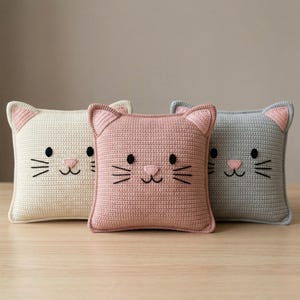 Peut inclure: Trois coussins carr&eacute;s au crochet, cr&egrave;me, rose et gris, chacun avec un motif de visage de chat. Les visages de chat ont des yeux et des moustaches brod&eacute;s en noir, avec des nez et des oreilles roses. Les coussins sont dispos&eacute;s sur une surface marron clair.
