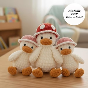 May include: Three handmade crochet duck toys. Each duck is white with an orange beak and feet. Two ducks wear pink hats, and one wears a red mushroom hat with white spots. The image includes the text "Instant PDF Download".