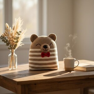May include: A handmade, crocheted bear tea cozy with a smiling face and a red bow tie. The cozy is beige with brown and white stripes. A cup of coffee and a vase of dried flowers sit on a wooden table.
