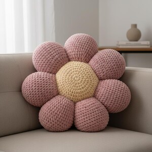 May include: A large, flower-shaped decorative pillow with a soft, textured appearance. The pillow features a light yellow center surrounded by eight pink petals. The pillow is made of a thick, knitted material and rests on a beige sofa.
