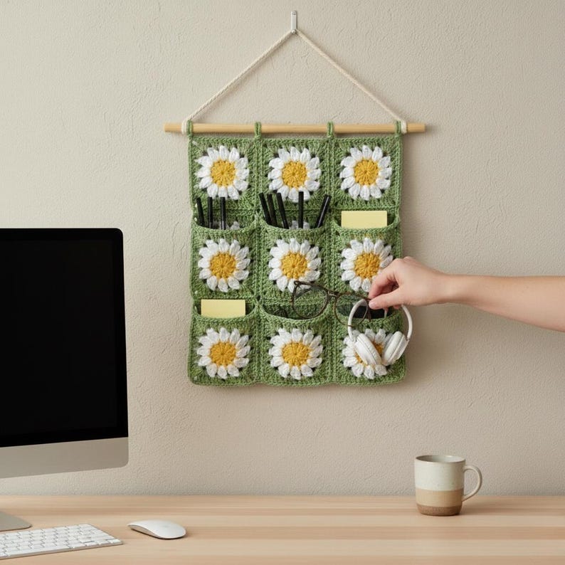 May include: A green crochet wall organizer with nine pockets, each adorned with a white and yellow daisy. The organizer hangs from a wooden dowel and a natural rope. The pockets hold pens, sticky notes, glasses, and headphones.