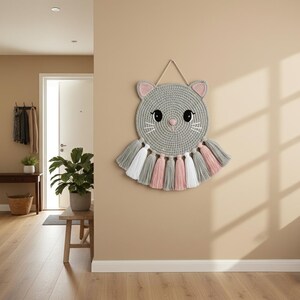 May include: A grey, crocheted cat wall hanging with pink and white details. The cat features a circular head with pink ears, black eyes, and white whiskers. Below the head are tassels in grey, pink, and white. It is suspended by a beige string.