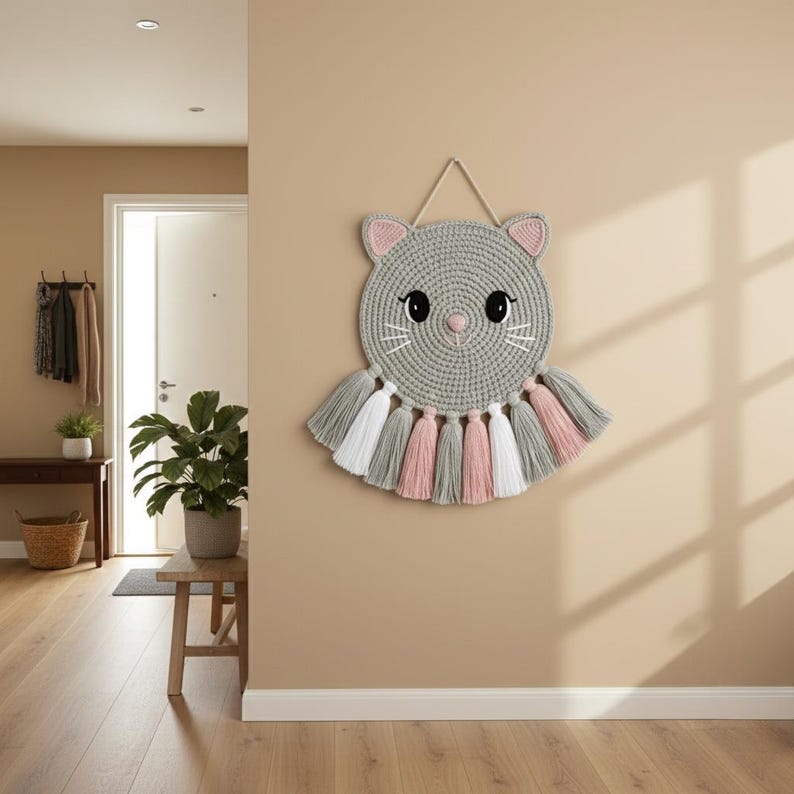 May include: A grey, crocheted cat wall hanging with pink and white details. The cat features a circular head with pink ears, black eyes, and white whiskers. Below the head are tassels in grey, pink, and white. It is suspended by a beige string.