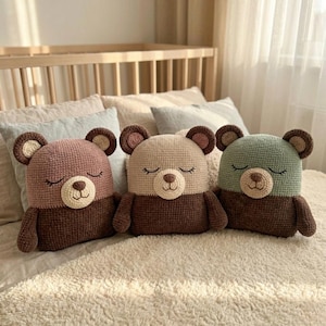 Könnte beinhalten: Drei handgefertigte Häkelbären-Kissen in Rosa, Beige und Grün. Jeder Bär hat eine braune Schnauze, Ohren und einen Unterkörper mit geschlossenen Augen. Die Kissen werden auf einem Bett mit einer neutralfarbenen Decke präsentiert.