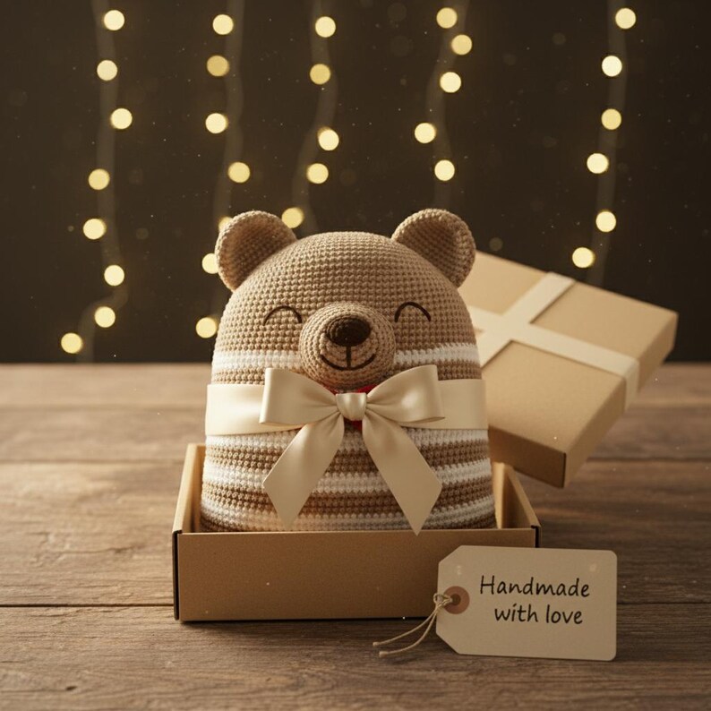 May include: A handmade crochet bear hat with a cream-colored bow and a red accent, presented in a cardboard box. The hat features brown and white stripes. A tag reads "Handmade with love."