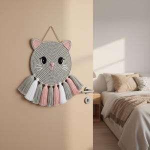 May include: A grey crocheted cat wall hanging with pink and white details. The cat has large black eyes, a pink nose, and whiskers. The hanging features tassels in grey, pink, and white. A decorative piece for a child's bedroom.