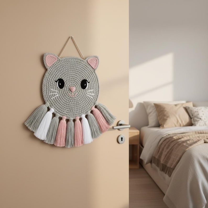 May include: A grey crocheted cat wall hanging with pink and white details. The cat has large black eyes, a pink nose, and whiskers. The hanging features tassels in grey, pink, and white. A decorative piece for a child's bedroom.