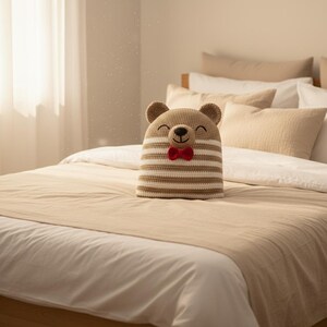 May include: A handmade, crocheted bear-shaped pillow with a smiling face and a red bow tie. The pillow features alternating horizontal stripes of beige and white. It is placed on a bed with neutral-toned bedding.