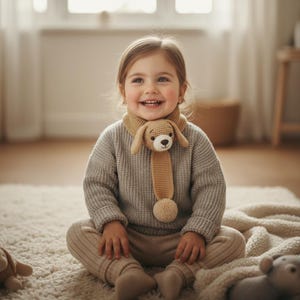May include: A young child wearing a tan crocheted dog scarf and a gray sweater sits on a white rug. The scarf features a dog's head and a dangling pom-pom. The child is smiling.