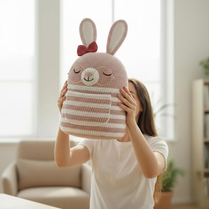 May include: A handmade, crochet bunny backpack in soft pink and white stripes. The bunny has large ears, a red bow, and round glasses. The backpack has a front pocket and is held by a person.