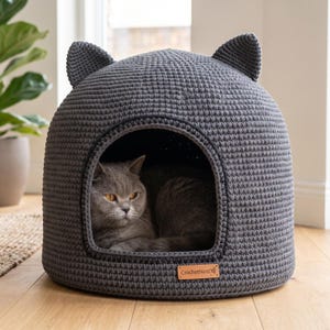 May include: A dark grey crocheted pet bed shaped like a cat head with two pointed ears on top. The bed features a wide front opening and a small rectangular tan leather tag with the text CrochetNest attached to the lower front edge.