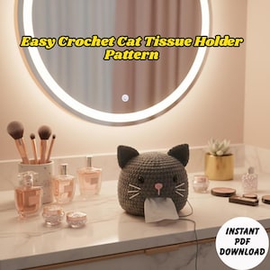 Op de afbeelding: Een grijze gehaakte katten-tissuehouder met witte snorharen en een roze neus, die tissues afgeeft. De tekst "Easy Crochet Cat Tissue Holder Pattern" wordt hierboven weergegeven. Op een wit aanrecht met schoonheidsproducten.