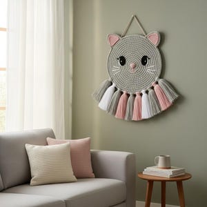 May include: A grey crochet cat wall hanging with pink and white details. The cat features large black eyes, a pink nose, and whiskers. Below the cat's face are grey, pink, and white tassels. The wall hanging is displayed on a green wall.