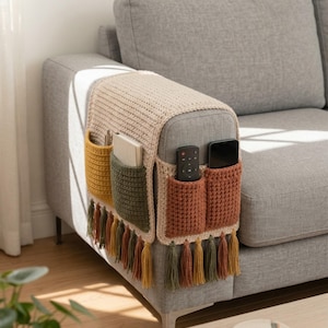 K&ouml;nnte beinhalten: Beige geh&auml;kelter Sofa-Arm-Organizer mit mehreren Taschen in Senfgelb, Olivgr&uuml;n und Rostfarben. Der Organizer hat dekorative Quasten in passenden Farben und h&auml;lt eine Fernbedienung, ein Telefon und ein Buch.