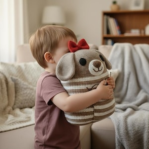 Könnte beinhalten: Ein handgefertigtes Häkelhundespielzeug in Beige- und Brauntönen mit einer roten Schleife. Der Hund hat einen gestreiften Körper, Schlappohren und ein lächelndes Gesicht. Das Spielzeug wird von einem Kind gehalten.