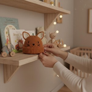 Pode incluir: Um gorro de beb&ecirc; laranja tricotado com orelhas de raposa e um desenho de rosto est&aacute; em uma prateleira de madeira. O chap&eacute;u est&aacute; ao lado de livros infantis e brinquedos de madeira. A prateleira faz parte da decora&ccedil;&atilde;o de um ber&ccedil;&aacute;rio.