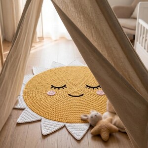 Puede incluir: Una alfombra de ganchillo amarilla y blanca con forma de sol sonriente con los ojos cerrados y mejillas rosas. La alfombra est&aacute; sobre un suelo de madera, con una estructura tipo tienda de campa&ntilde;a beige al fondo. Un peluche est&aacute; al lado de la alfombra.