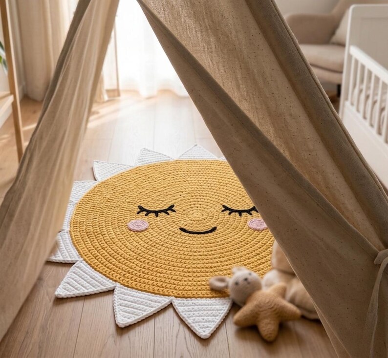 Puede incluir: Una alfombra de ganchillo amarilla y blanca con forma de sol sonriente con los ojos cerrados y mejillas rosas. La alfombra est&aacute; sobre un suelo de madera, con una estructura tipo tienda de campa&ntilde;a beige al fondo. Un peluche est&aacute; al lado de la alfombra.