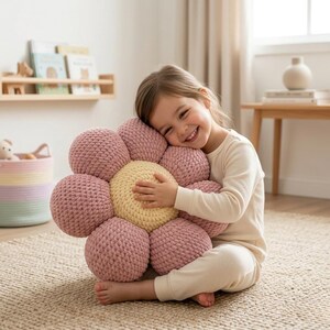 May include: A large, handmade flower-shaped pillow in shades of pink and yellow. The pillow is made of a soft, textured material and has a round center with multiple petal-like spheres surrounding it. The pillow is on a neutral-colored rug.