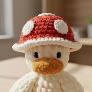 May include: A plush white duck wearing a red and white crochet mushroom hat. The hat has white spots and a white brim. The duck has a yellow-orange beak and black eyes. The toy is made of soft, textured yarn.