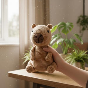 Pu&ograve; includere: Un peluche di capibara all'uncinetto fatto a mano, nei toni del beige e del marrone. L'animale di peluche ha una testa rotonda con piccole orecchie e un muso marrone scuro, ed &egrave; seduto con le braccia e le gambe distese. Il giocattolo &egrave; tenuto davanti a un tavolo di legno, con piante sullo sfondo.