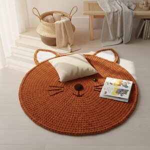 May include: A round, rust-colored rug shaped like a fox's face, with white-tipped ears and embroidered details. A small pillow and a book rest on the rug. The rug is made of a thick, textured material.