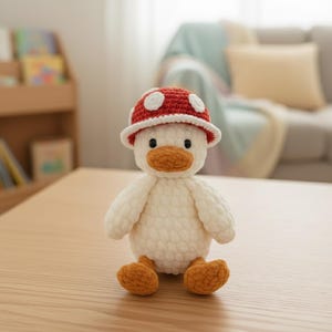 May include: A handmade white crochet duck wearing a red mushroom hat with white spots. The duck has an orange beak and feet. The plush toy is sitting on a light brown wooden surface.