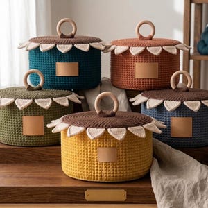 May include: Five colorful, crocheted storage baskets with lids. Each basket is a different color: teal, orange, olive green, yellow, and navy blue. The lids are brown with a scalloped edge and a wooden handle. Each basket has a small, square, tan leather tag.