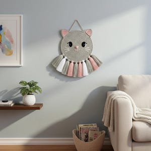 May include: A circular, grey crocheted cat wall decoration with pink ears and nose, black eyes, and white whiskers. It features a fringe of grey, white, and pink tassels. The cat is suspended by a grey cord, a charming addition to any room.