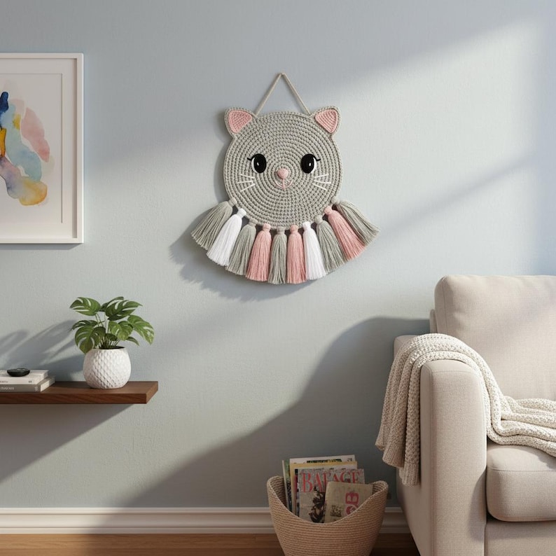 May include: A circular, grey crocheted cat wall decoration with pink ears and nose, black eyes, and white whiskers. It features a fringe of grey, white, and pink tassels. The cat is suspended by a grey cord, a charming addition to any room.