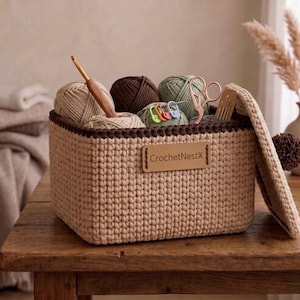 May include: A beige crochet basket filled with yarn balls in various colors, a crochet hook, scissors, and stitch markers. The basket has a brown trim and a leather tag that reads "CrochetNestX". The basket is on a wooden table.