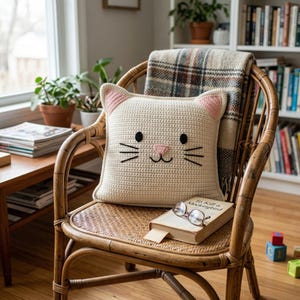 Peut inclure: Un coussin en crochet cr&egrave;me en forme de chat, avec des oreilles roses et un visage cousu, est pos&eacute; sur une chaise en osier. Une couverture &agrave; carreaux est drap&eacute;e sur la chaise, et un livre avec des lunettes est pos&eacute; en dessous. La sc&egrave;ne se d&eacute;roule dans une pi&egrave;ce chaleureuse et bien &eacute;clair&eacute;e.